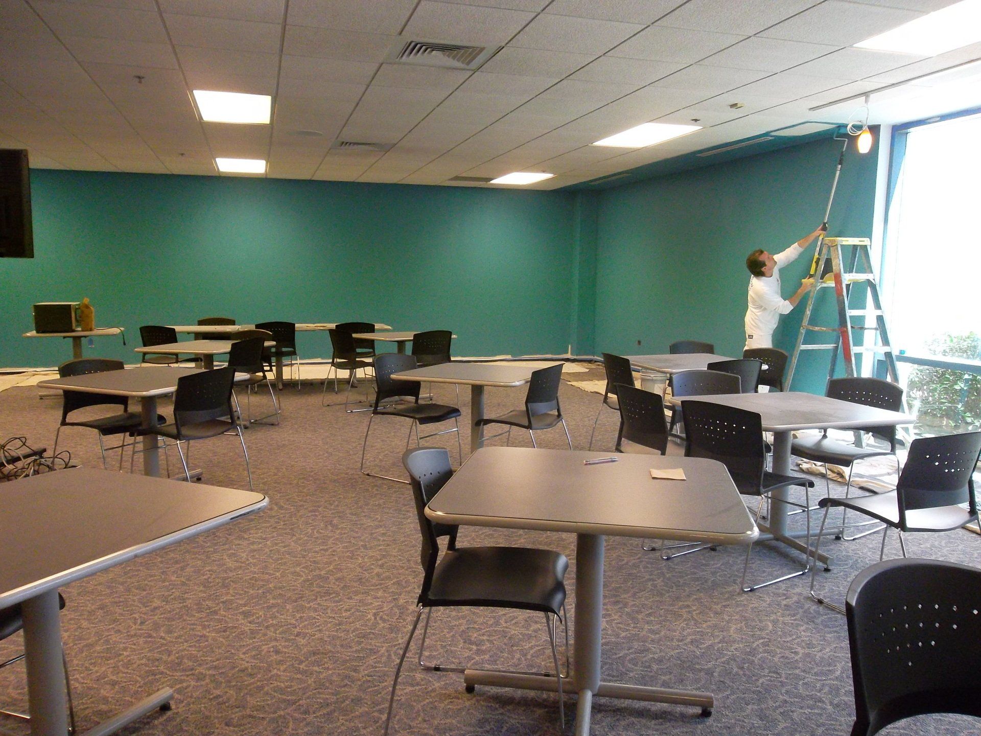 A man is painting a wall in a room with tables and chairs