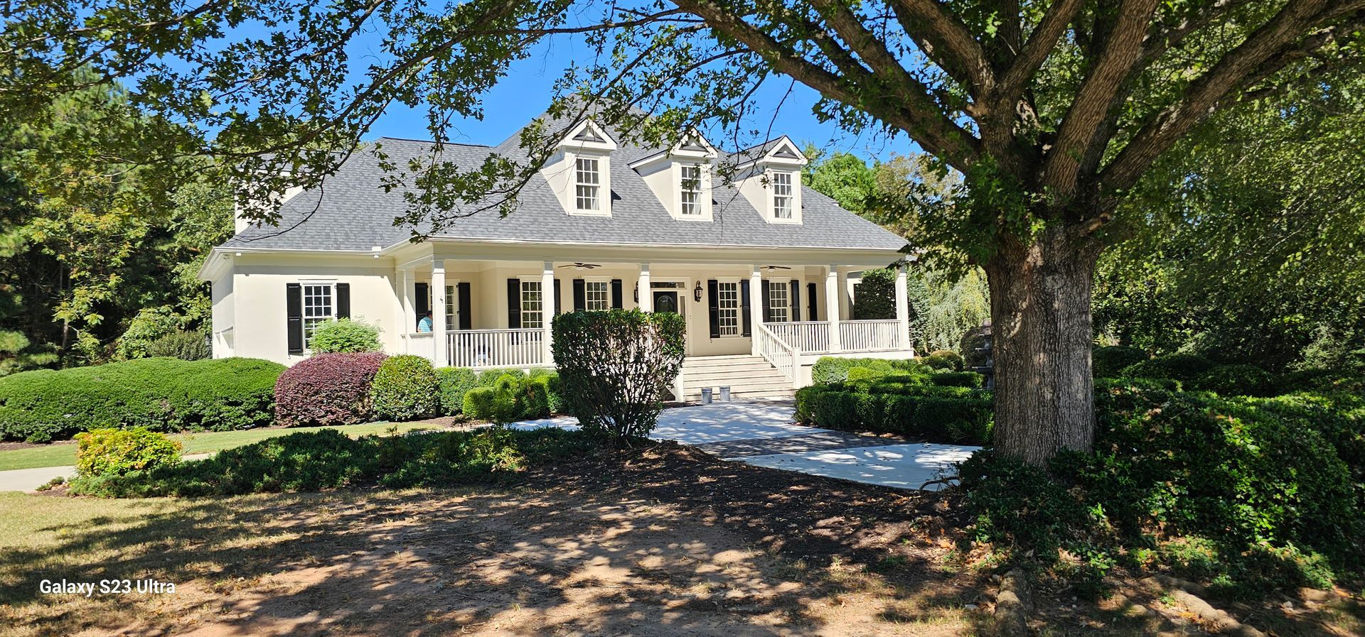 A large white house with a gray roof is surrounded by trees and bushes.