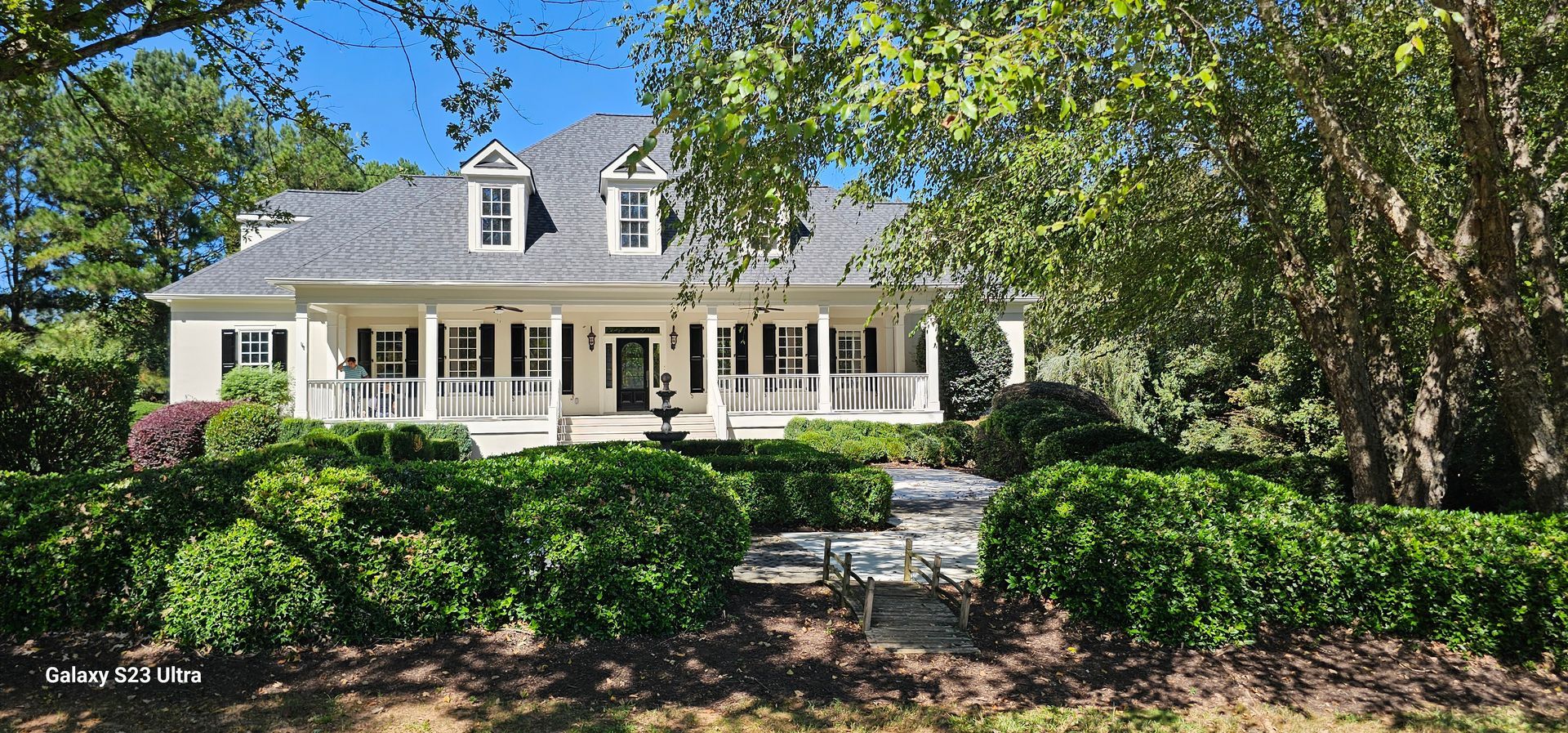 A large white house with a gray roof is surrounded by trees and bushes.