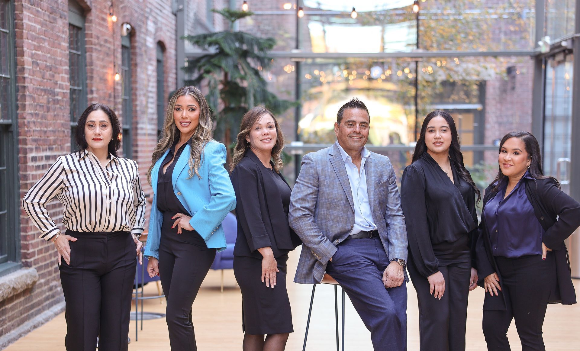 Six people in business attire pose inside a brick-walled building with large windows.