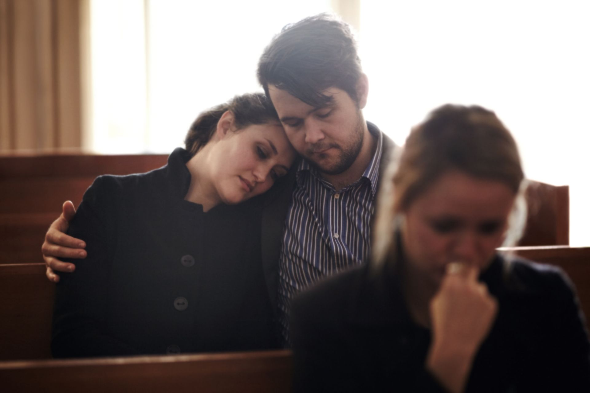 People comforting each other at a funeral; woman rests head on man's shoulder. Another woman nearby appears sad.