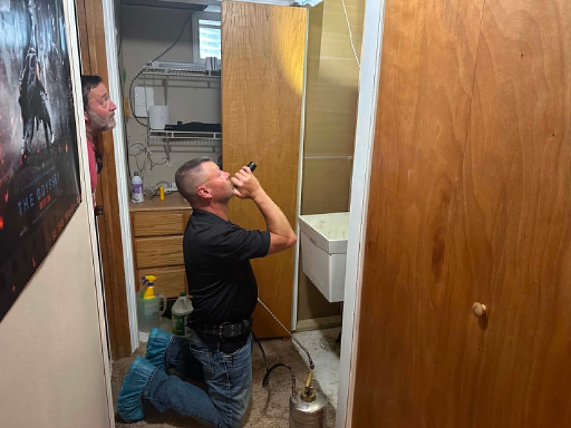 Man kneels on a bathroom floor while working on bug control in a narrow, wood-paneled space.