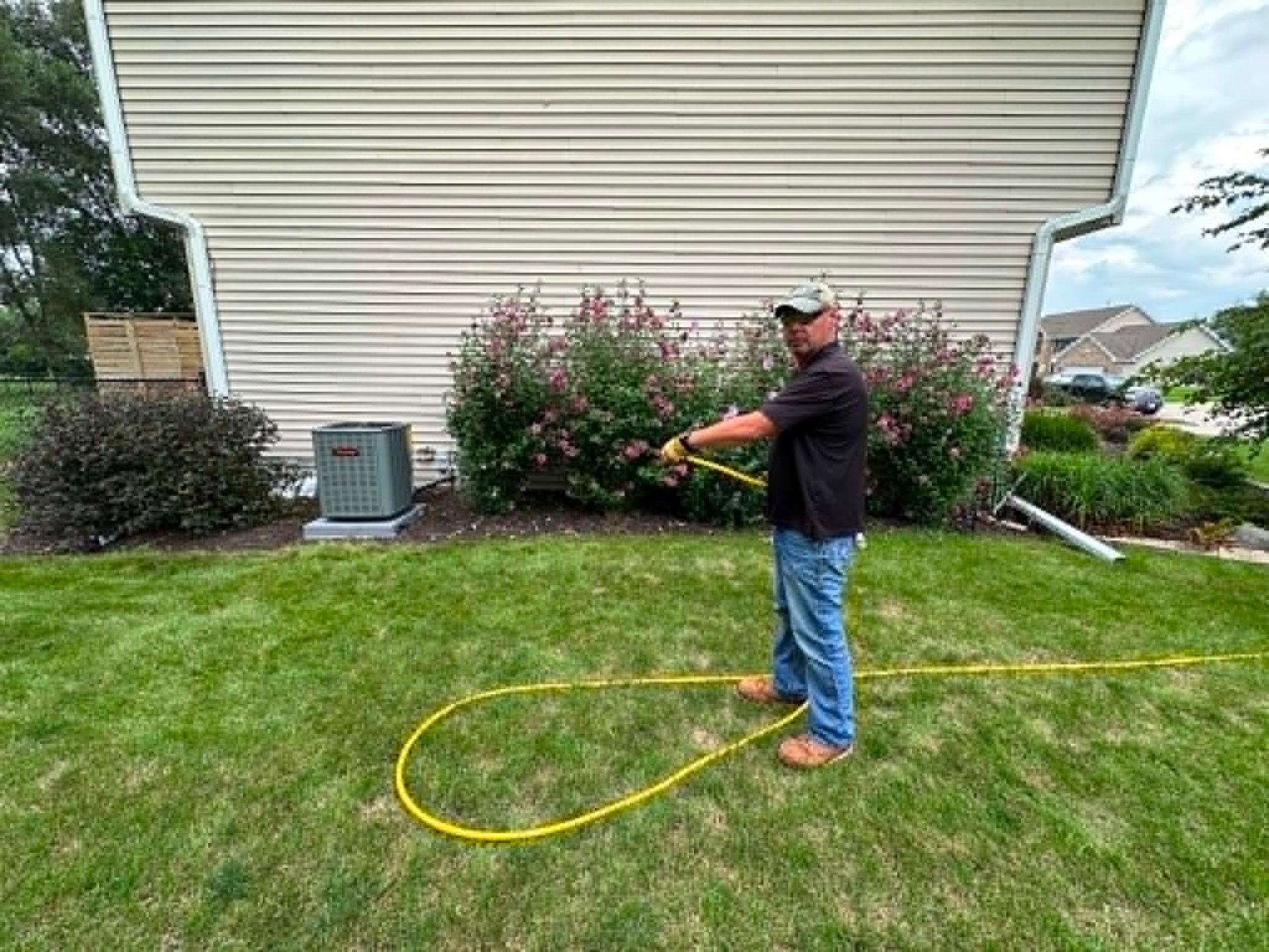 Man spraying lawn with a hose near a house and shrubs, yellow hose on green grass.