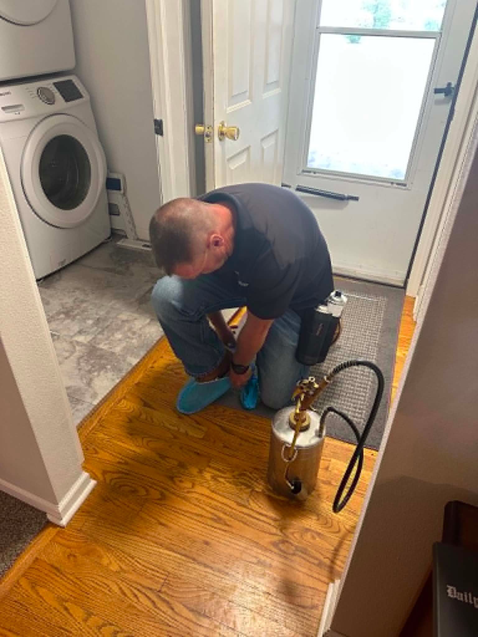 Technician kneeling on hardwood floor doing bug control near a laundry room door.