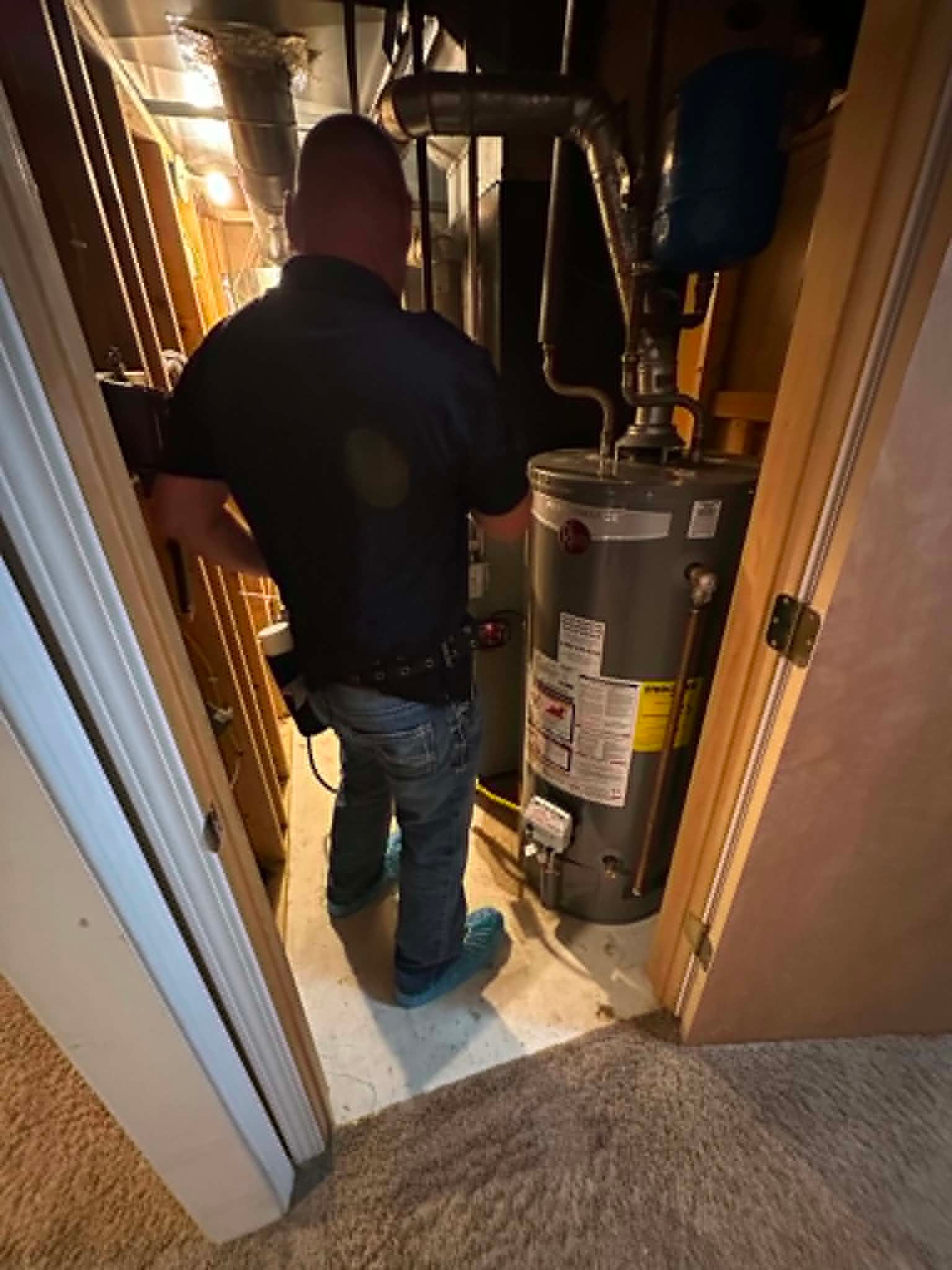 Person standing in a narrow basement hallway beside a water heater and utility pipes doing bug control.