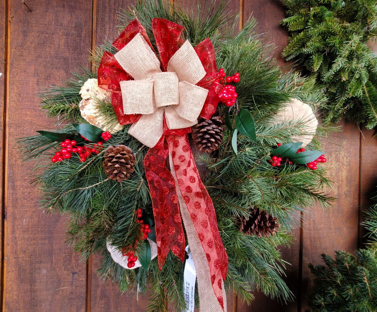 Christmas wreath with red and beige bows, pinecones, and berries, against a wooden background.