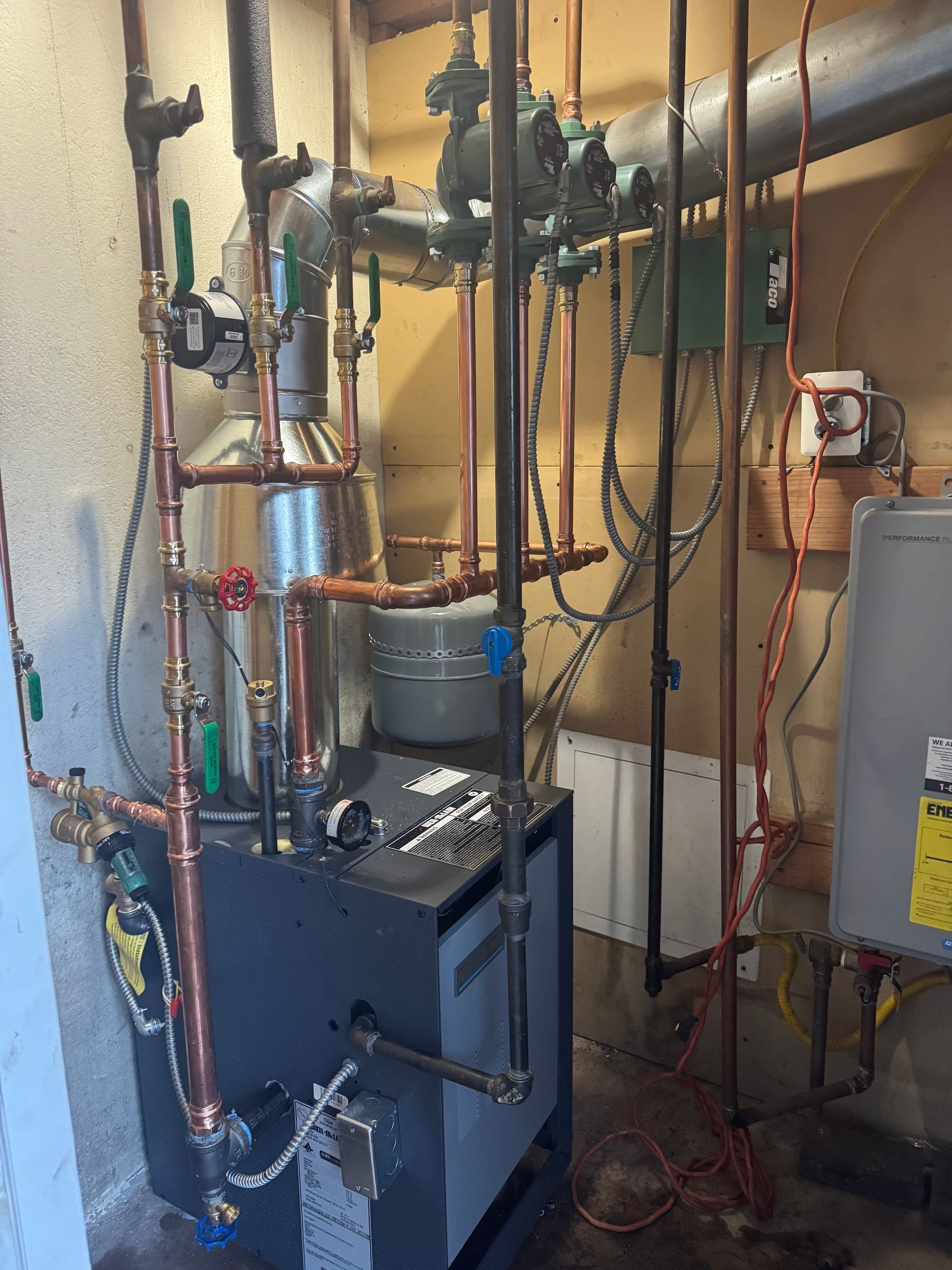 Boiler with copper pipes and various valves in a utility room. Grey and brown tones.