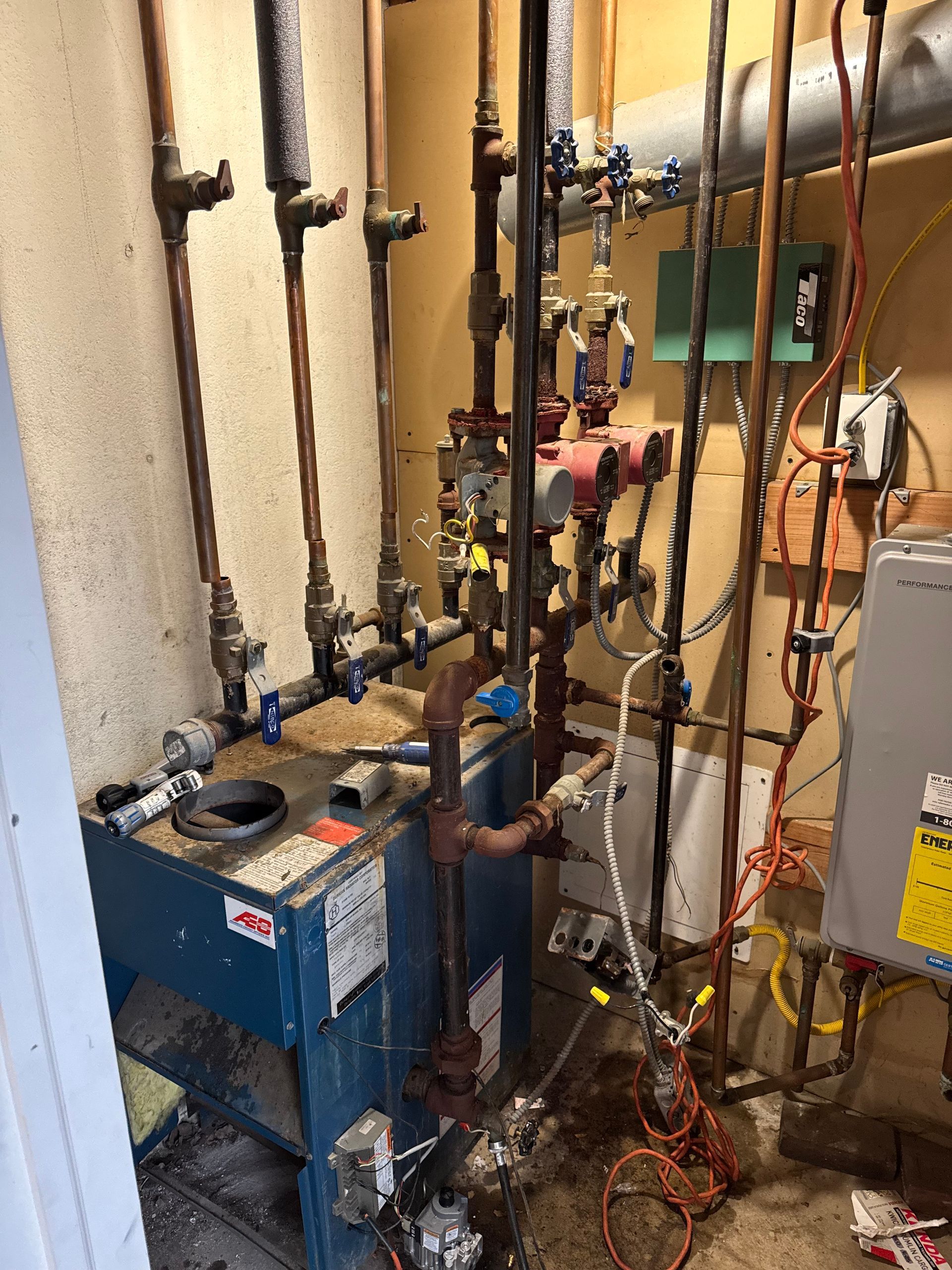 Boiler system with pipes and valves in a utility room. Blue boiler, copper and black pipes, and wires against a beige wall.