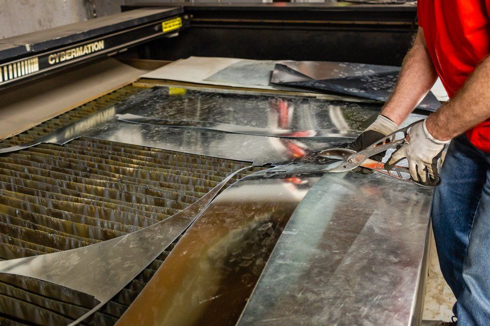 A person in red shirt and gloves cutting sheet metal with shears near a laser cutter.