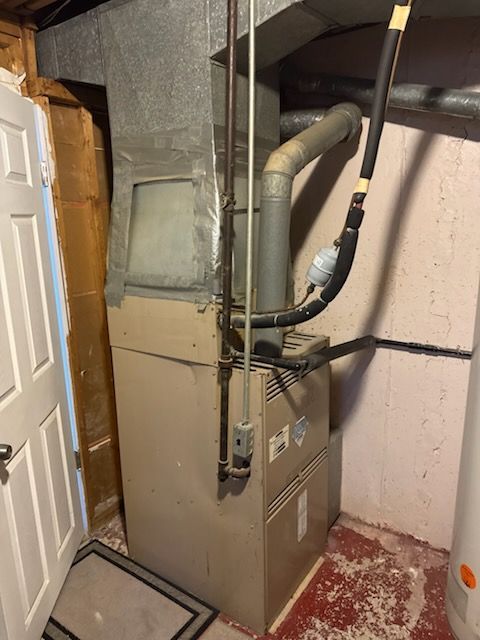 Furnace in a basement; gray metal, pipes, and ductwork. White door and red concrete floor.