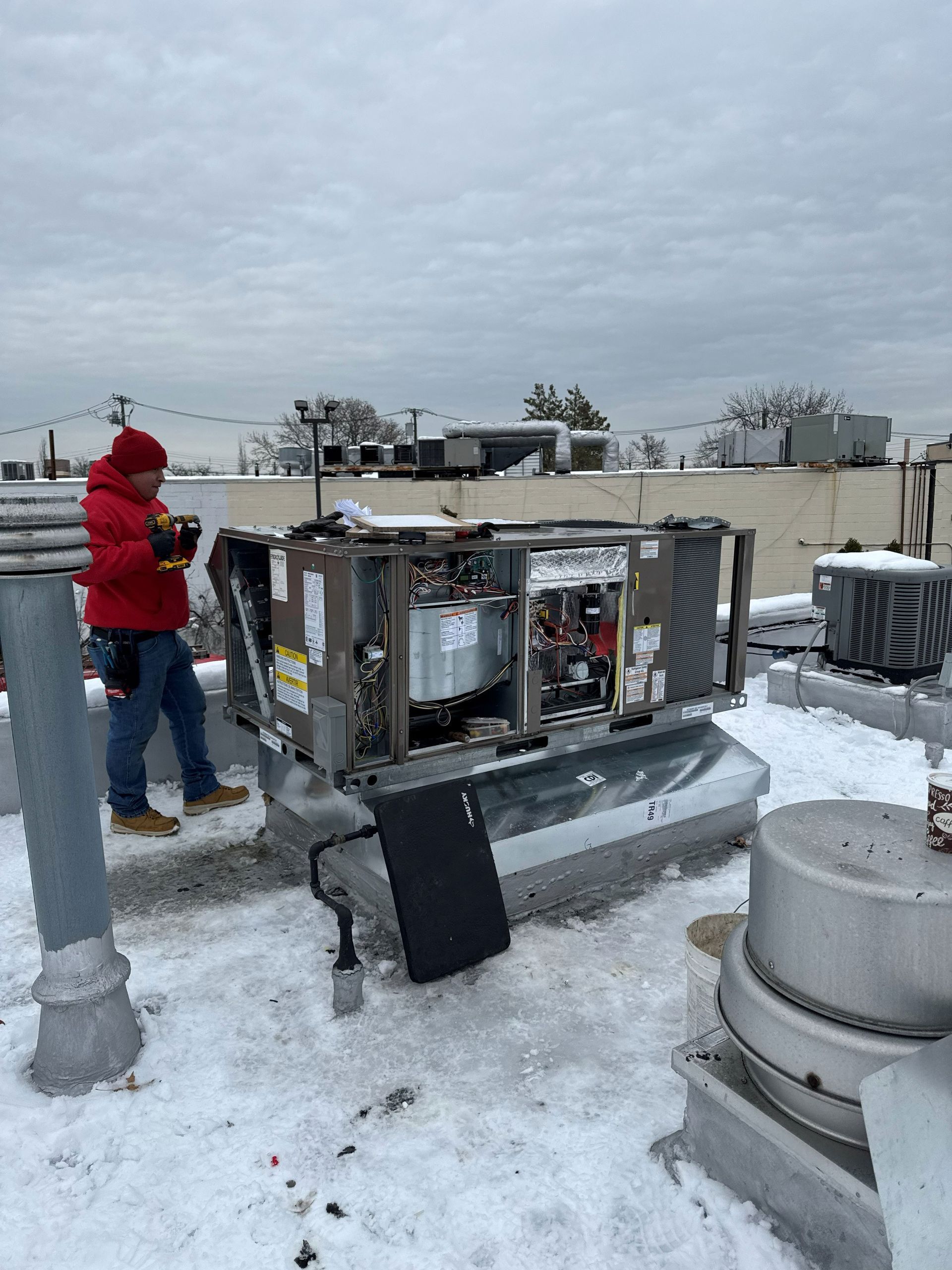 A person in a red jacket works on rooftop HVAC unit covered in snow on a cloudy day.