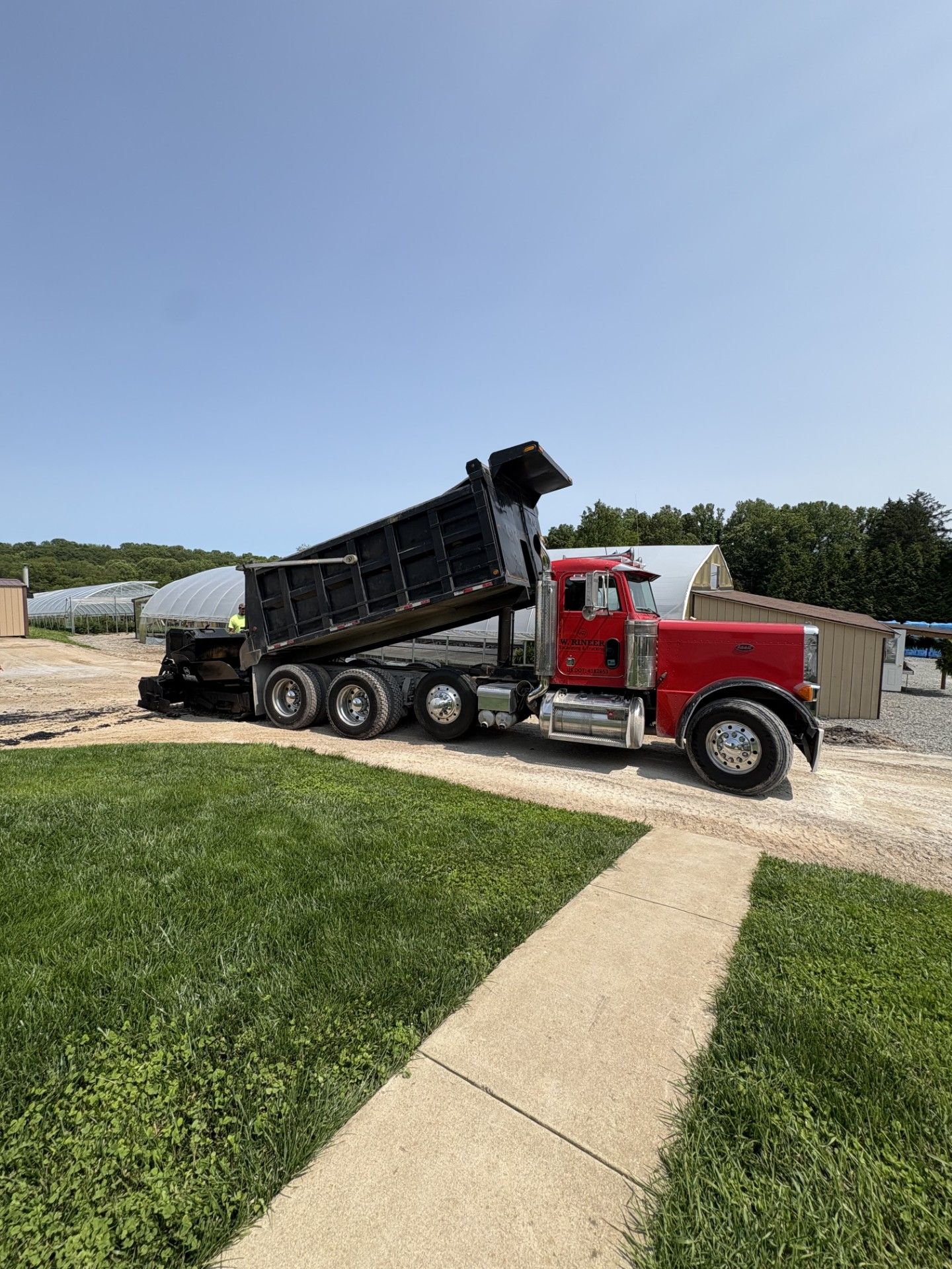 Red dump truck unloading on a dirt path near a green grassy area under a blue sky.