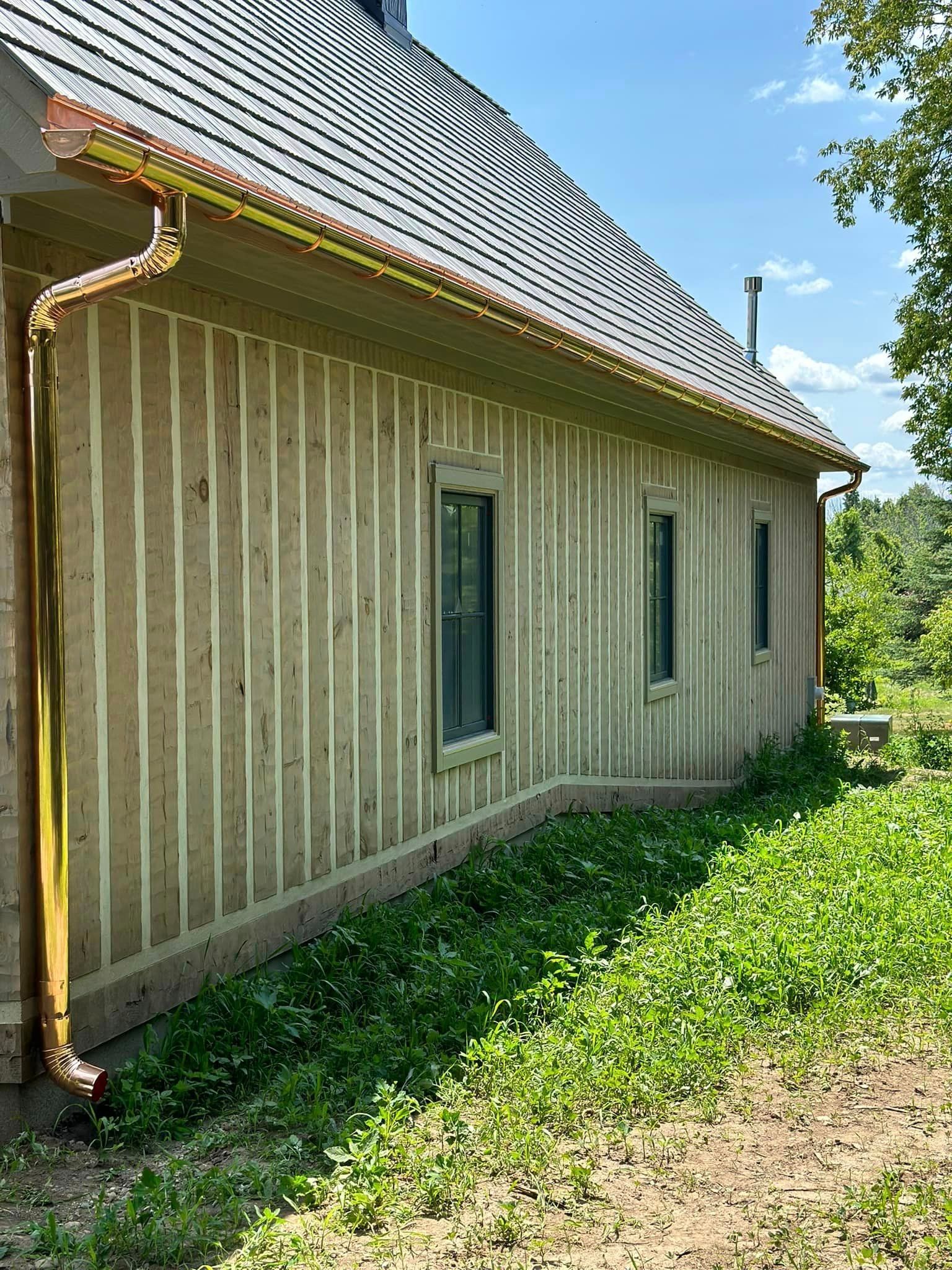 Side view of a house with wooden siding, copper gutters, and windows, overgrown with greenery.