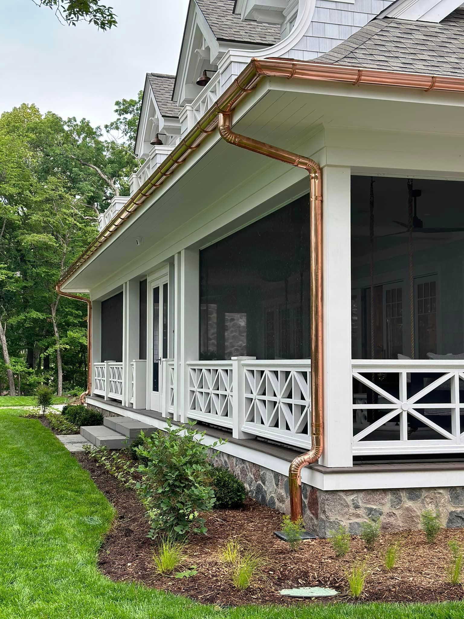 Copper-trimmed porch with white railings, a screened-in area, and a stone foundation. Lush green grass and landscaping.