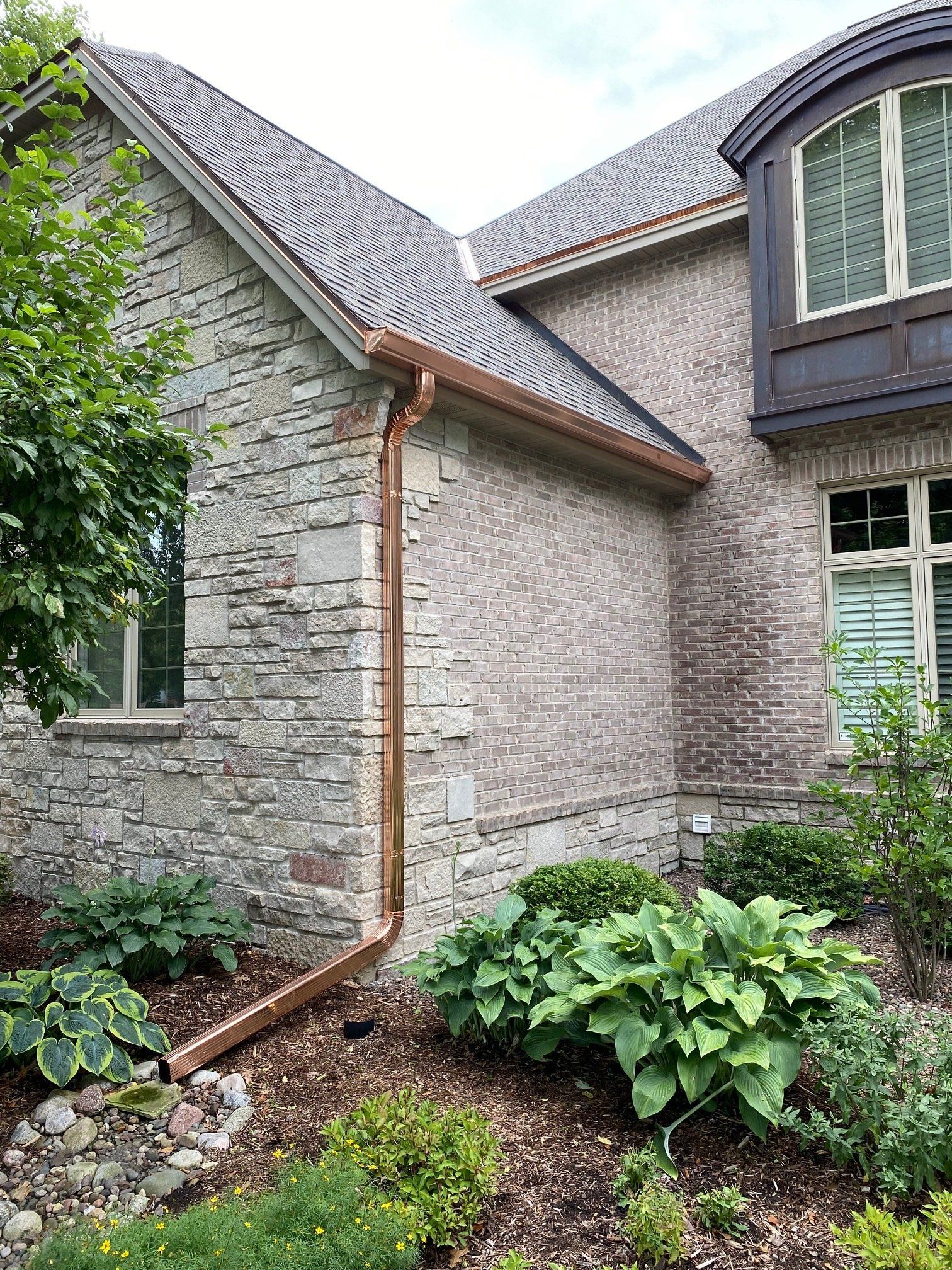 Copper gutters on a stone and brick house with landscaping and a dark shingle roof.