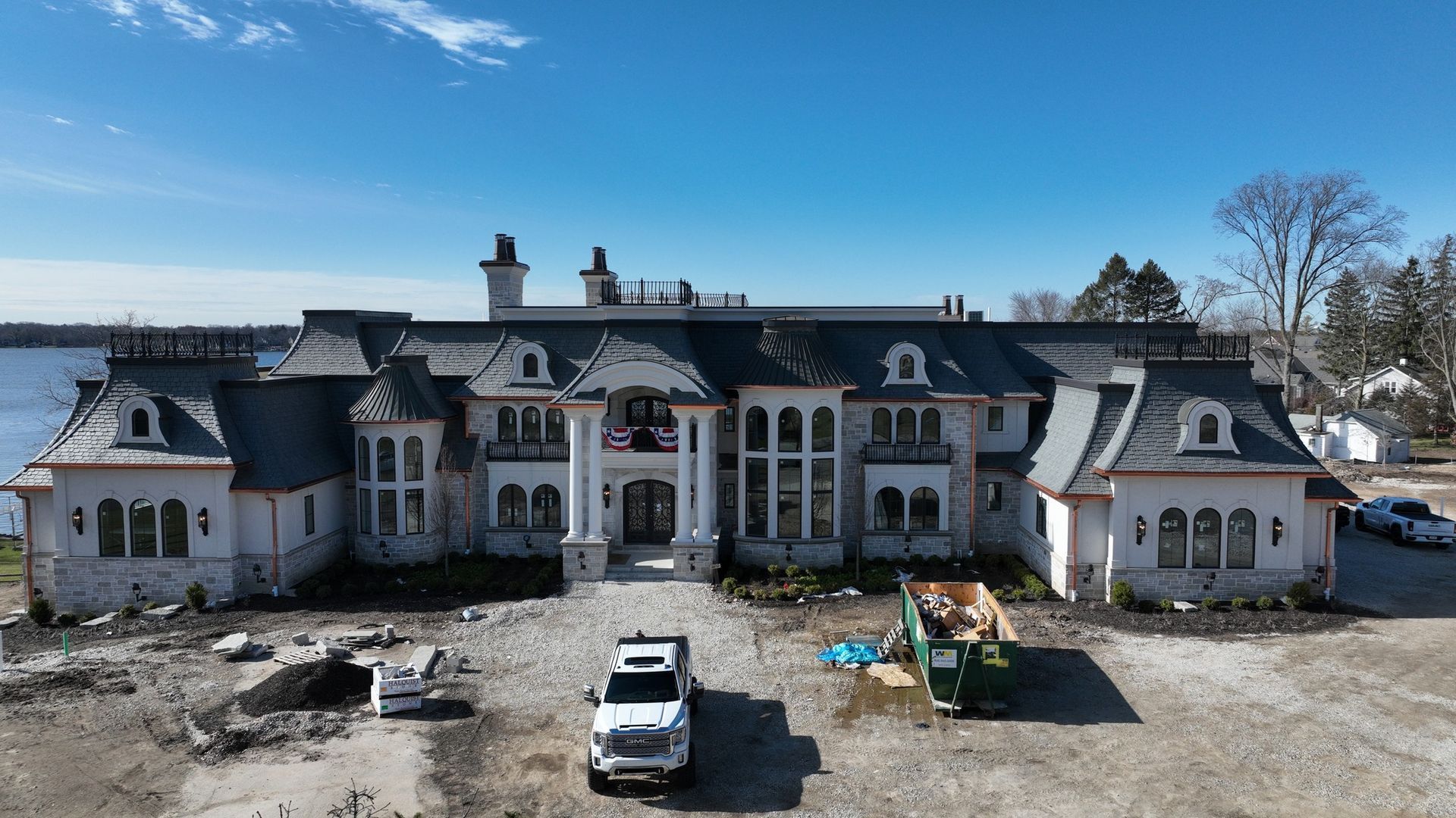 Large stone mansion with gray roof and white columns, under construction, with trucks and debris.