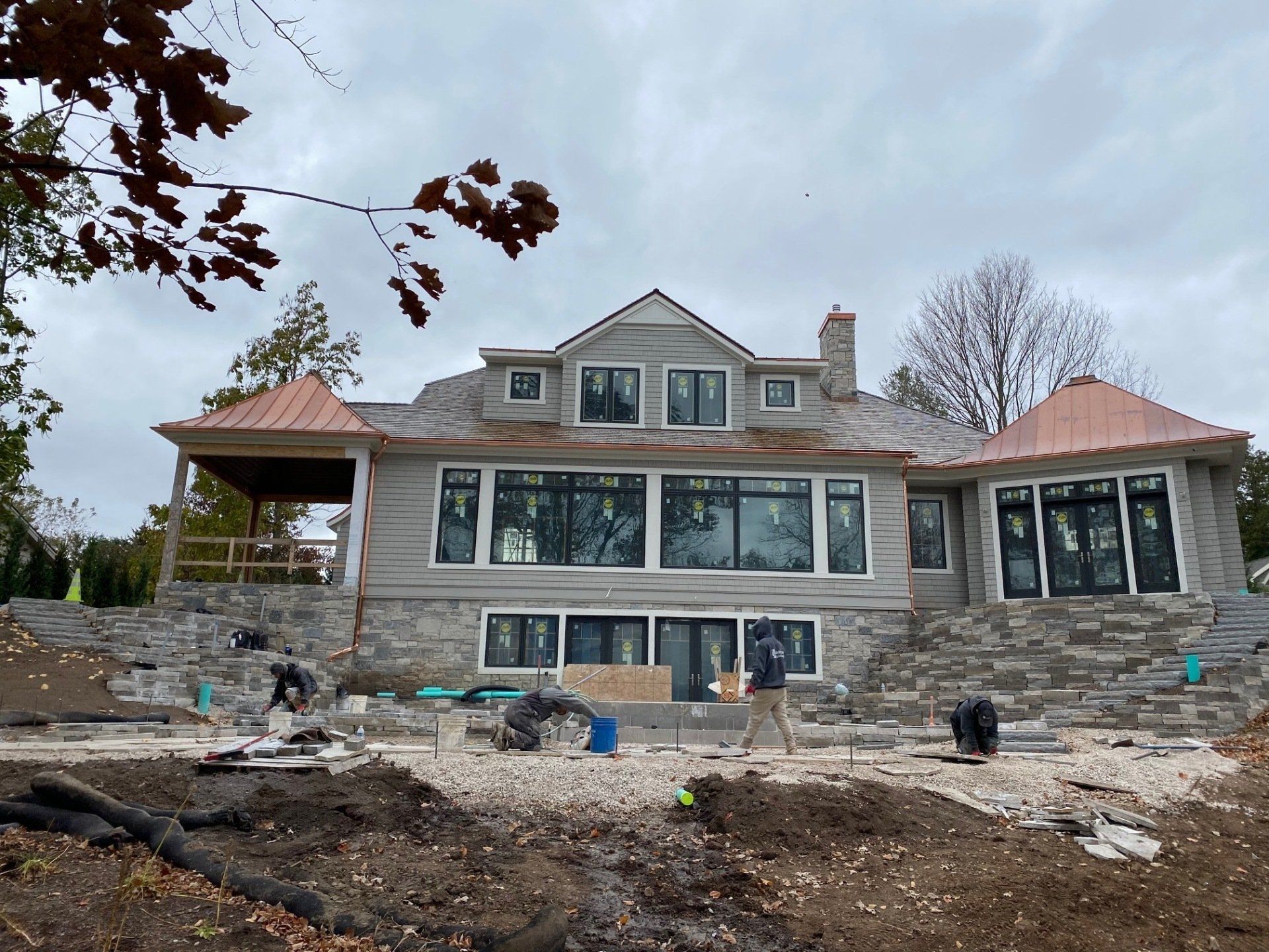 Large house under construction, gray siding, copper roof accents, large windows, workers on the lower level, cloudy sky.