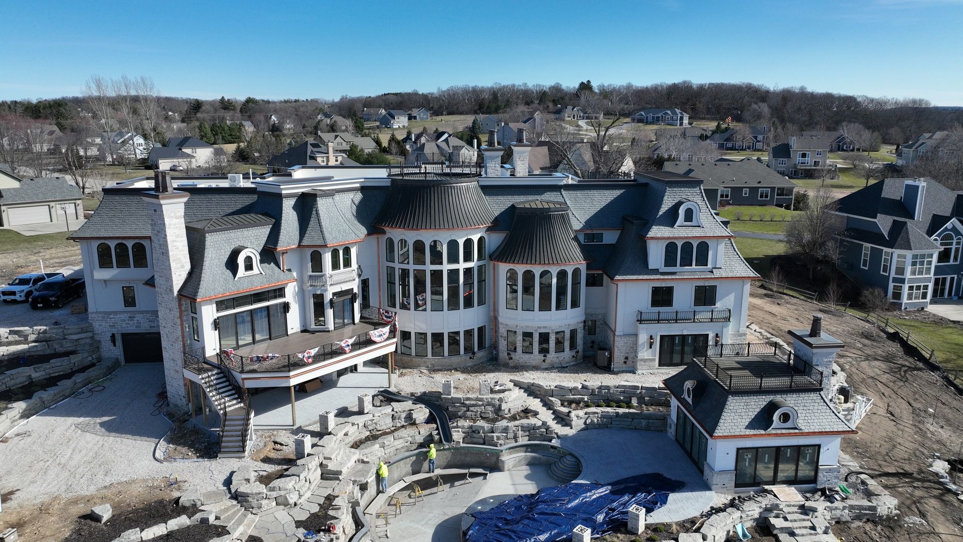 Large white mansion under construction with tiered stonework and a small pool on a hillside, overlooking a town.