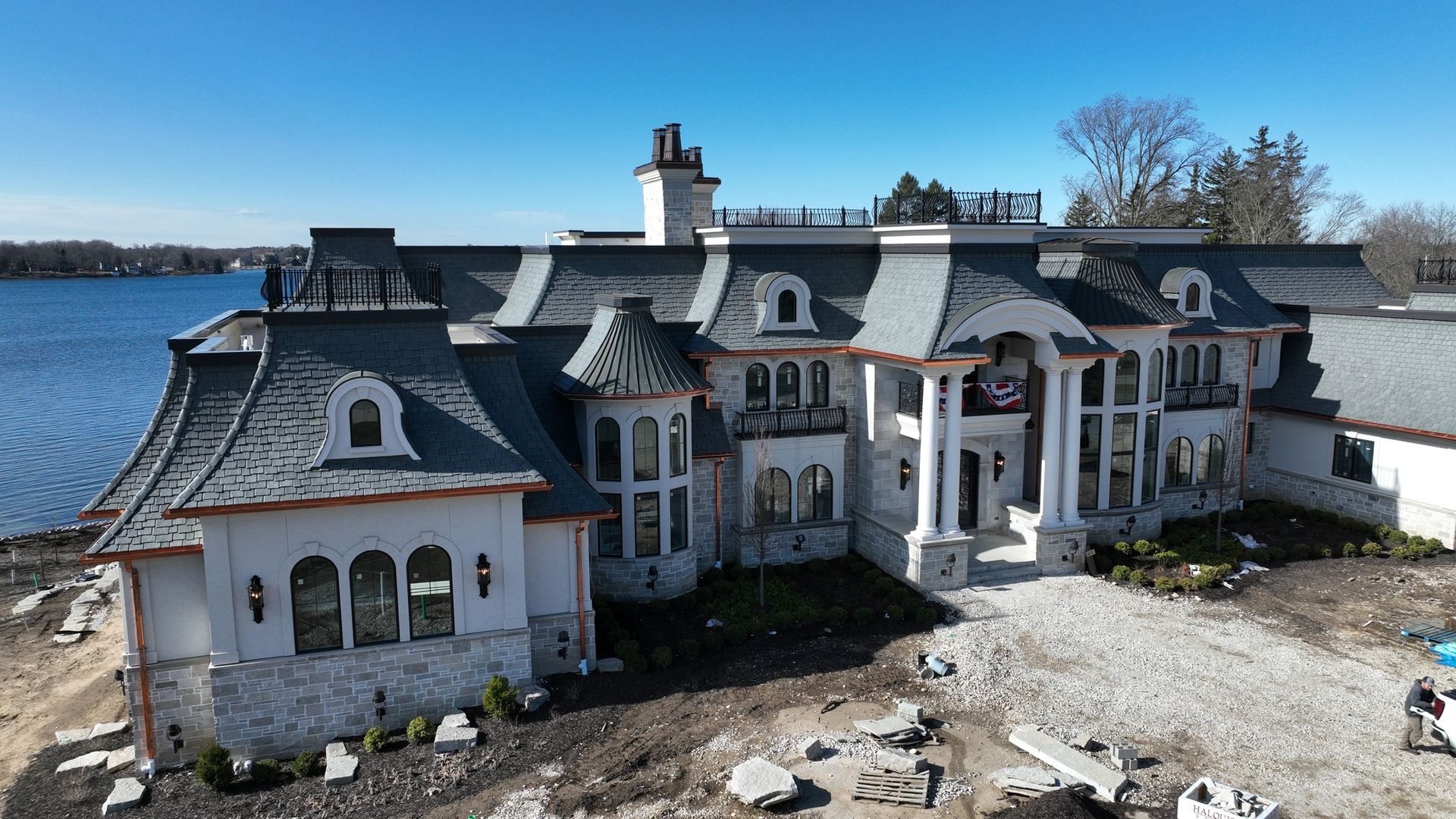 Large stone mansion with grey roof, columns, and lake view.