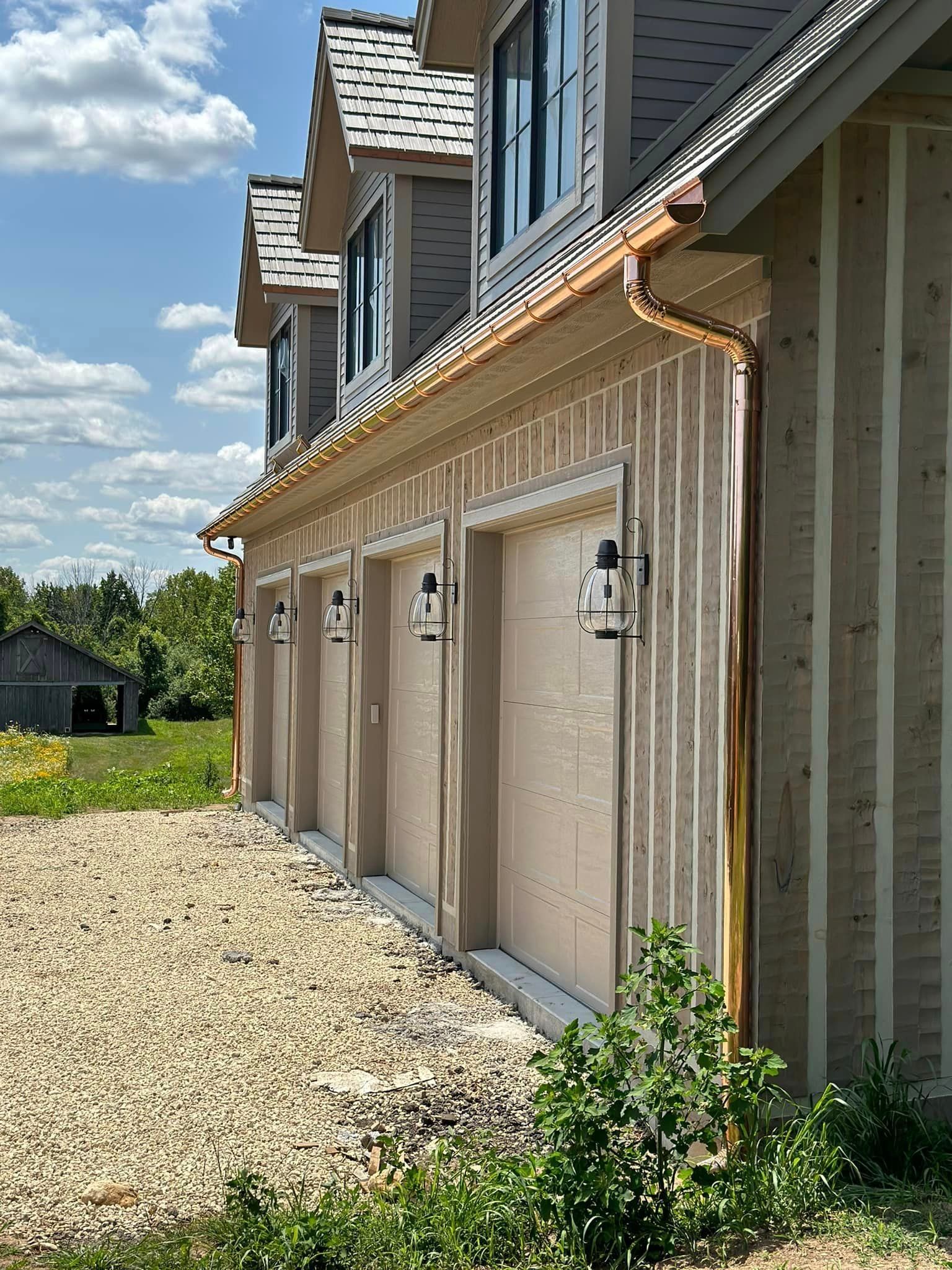 Side view of a garage with copper gutters and three overhead doors, with gravel and greenery in front.