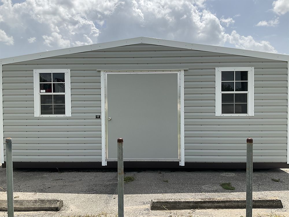 Small gray utility building with white trim, centered door, two windows, and a gravel lot in front