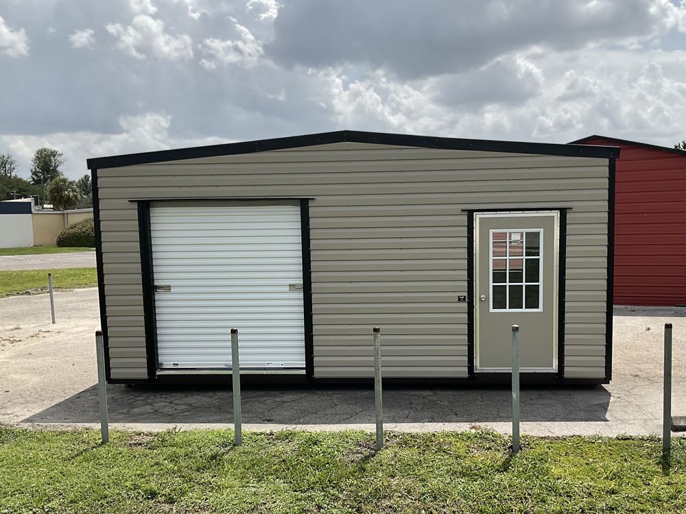 Small beige portable building with white roll-up door and side entry door behind metal posts on a lot