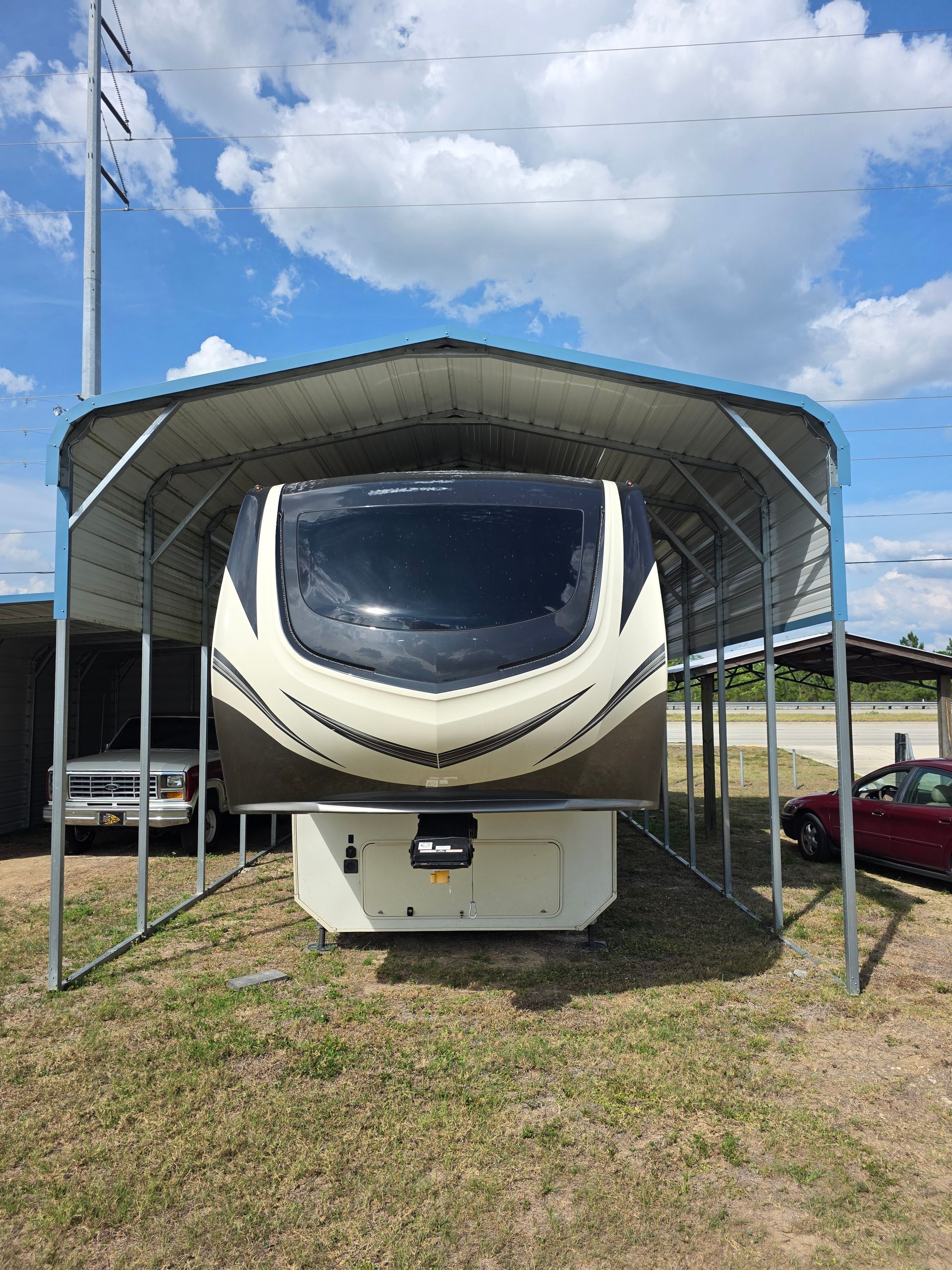 White RV parked under a metal canopy on grass, viewed from the front under a blue cloudy sky.