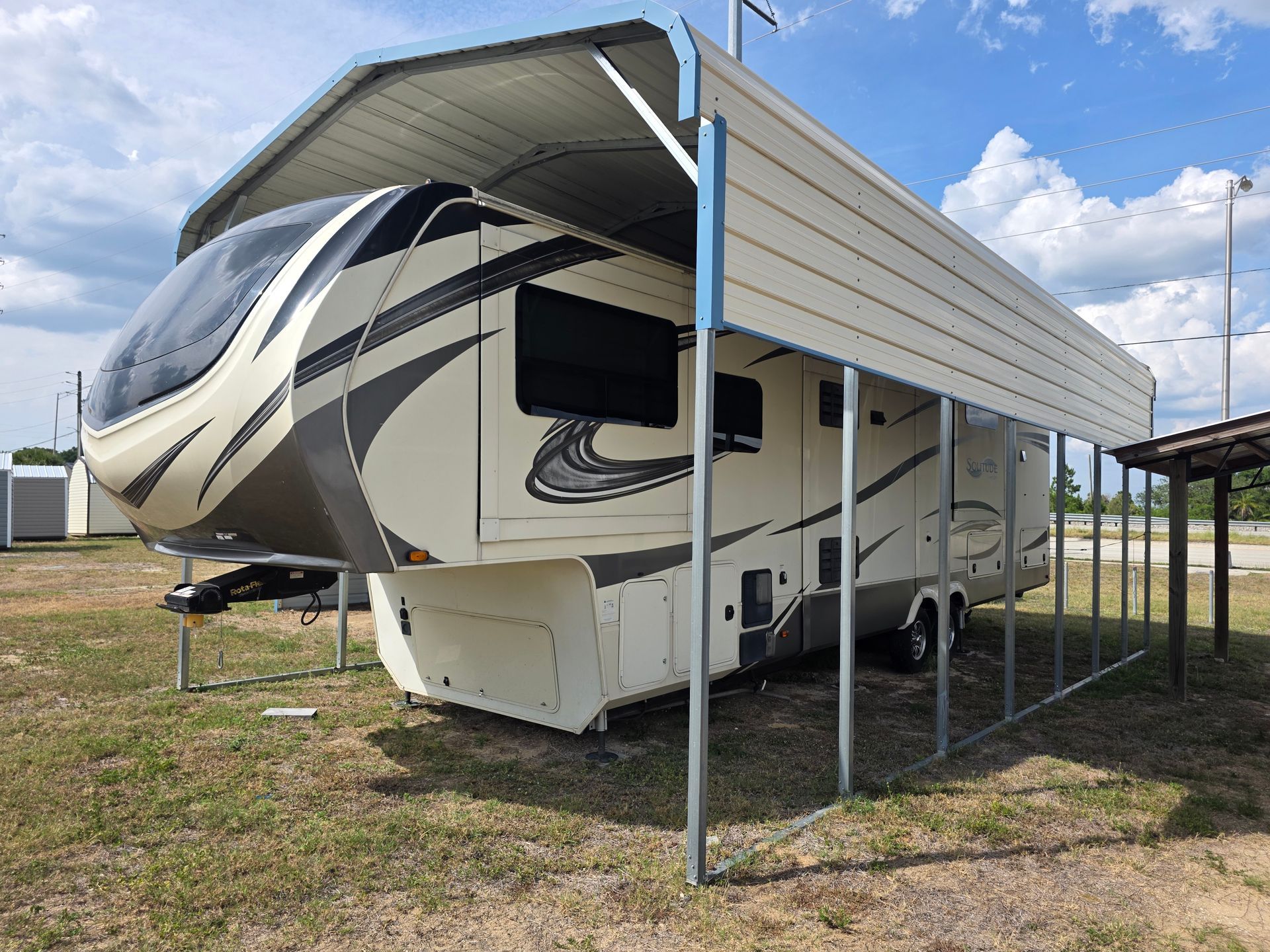 Large fifth-wheel RV trailer parked on grass under a metal carport, with a striped beige and black exterior.