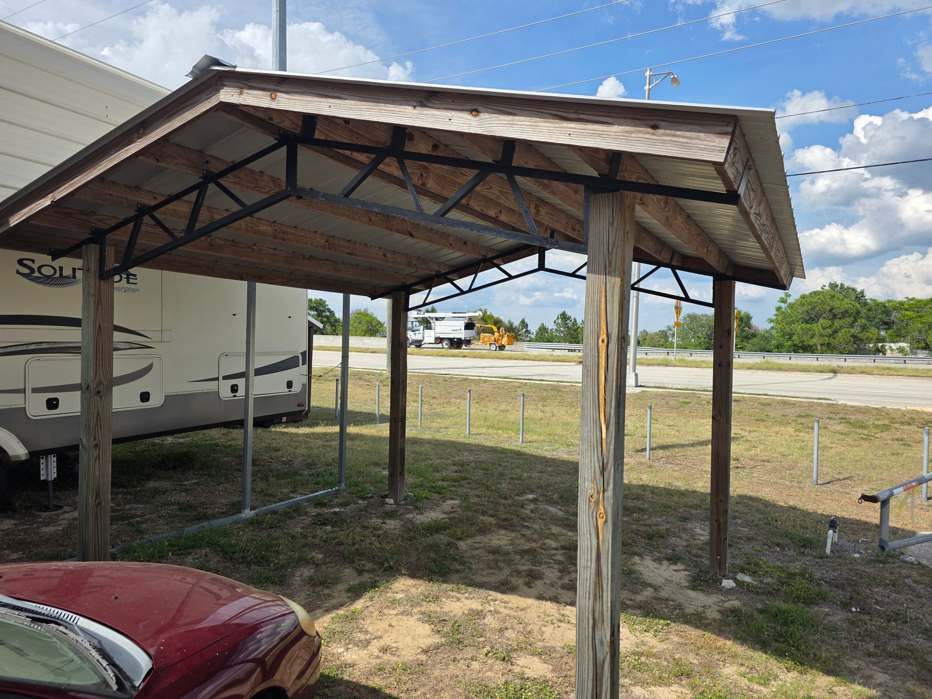 Rustic metal carport with a weathered roof beside a red car and RV in a grassy lot under a blue sky