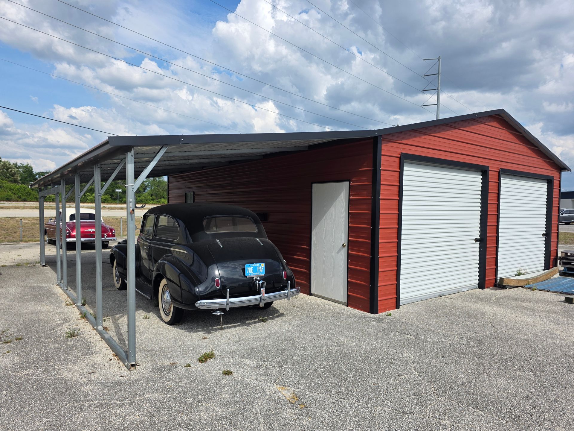 Black Volkswagen parked under a metal carport beside a red garage with white roll-up doors on gravel.