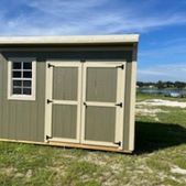 Small tan shed with white-trimmed windows and double doors on a grassy field under a blue sky
