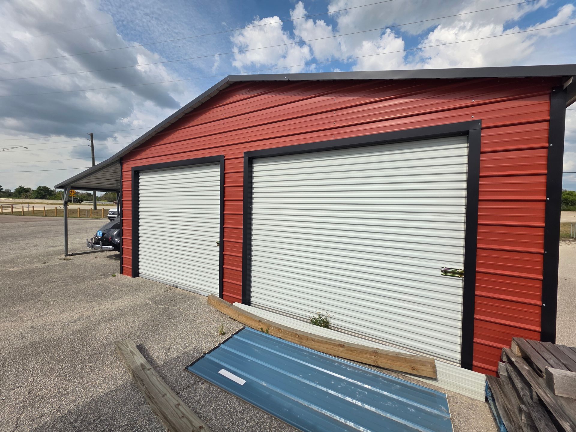 Red metal storage building with two white roll-up doors on a gravel lot under a cloudy sky
