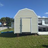 Beige shed with open door on a grassy lawn beside a parked RV under a blue sky.