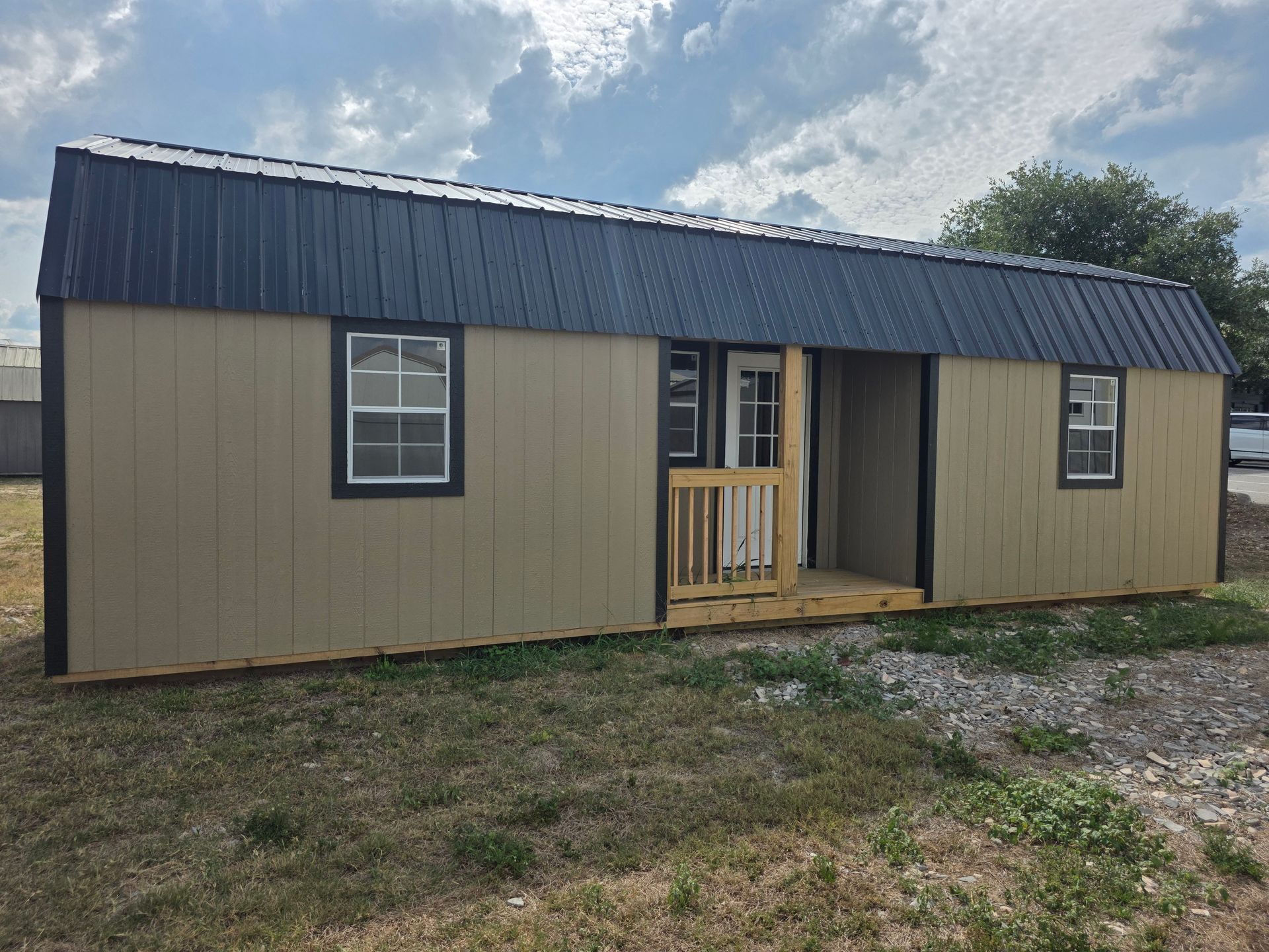 Small tan mobile home with dark roof and porch, set on a grassy lot under a cloudy sky.