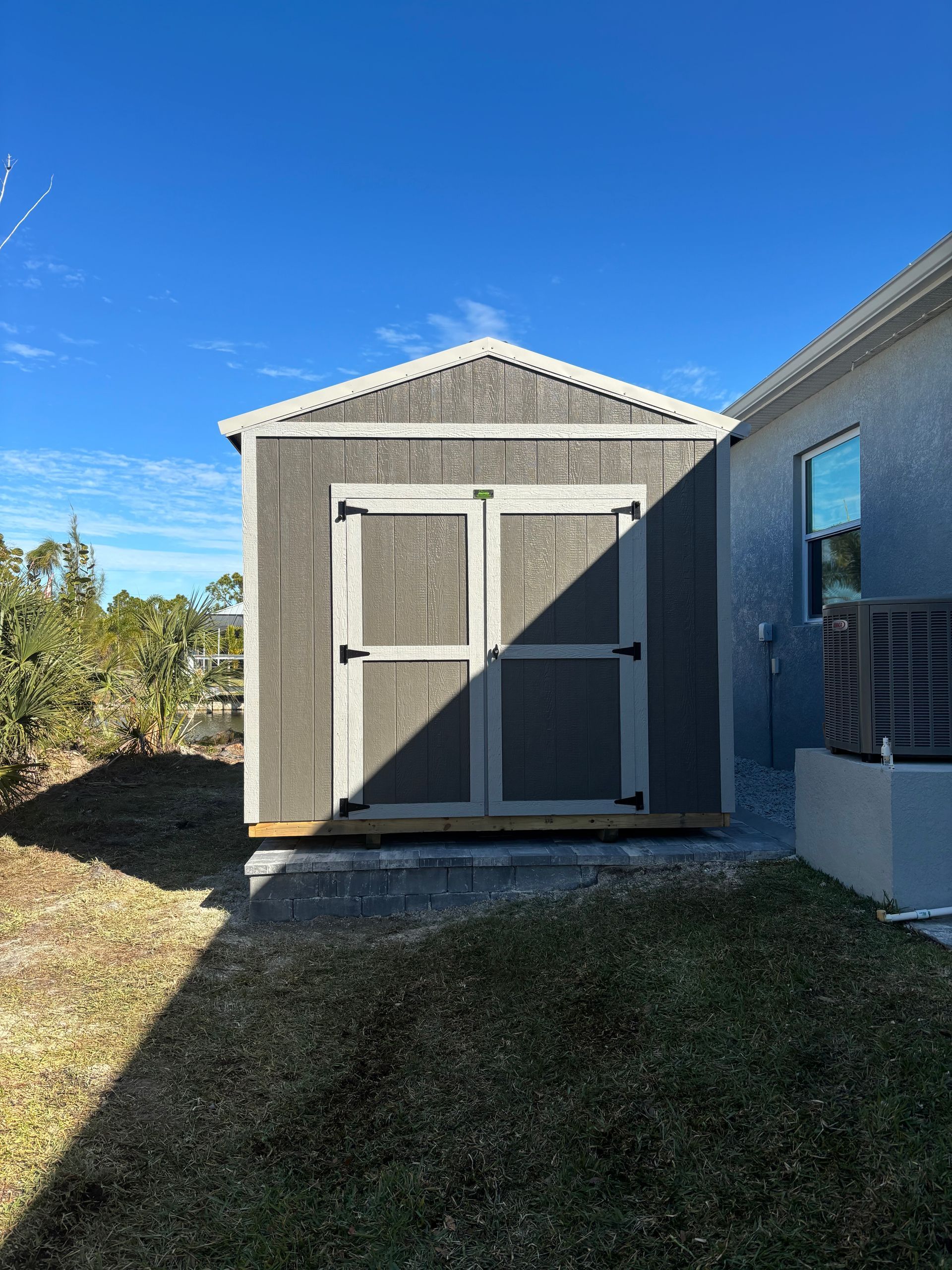 Small gray storage shed beside a house, with double doors, in a sunny yard