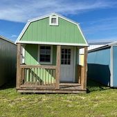 Small green shed with a porch and white trim in a grassy yard under a blue sky