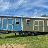 Two tiny mobile cabins, blue and gray, parked on a trailer in a grassy field under a blue sky.