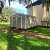 White storage shed in a grassy yard beside a beige building, with trees casting shade.