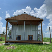 Small wooden building with wire mesh walls and a peaked roof on a grassy lot under a cloudy sky