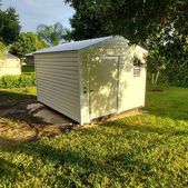 Small white storage shed with a slanted roof on a grassy lawn, surrounded by trees.