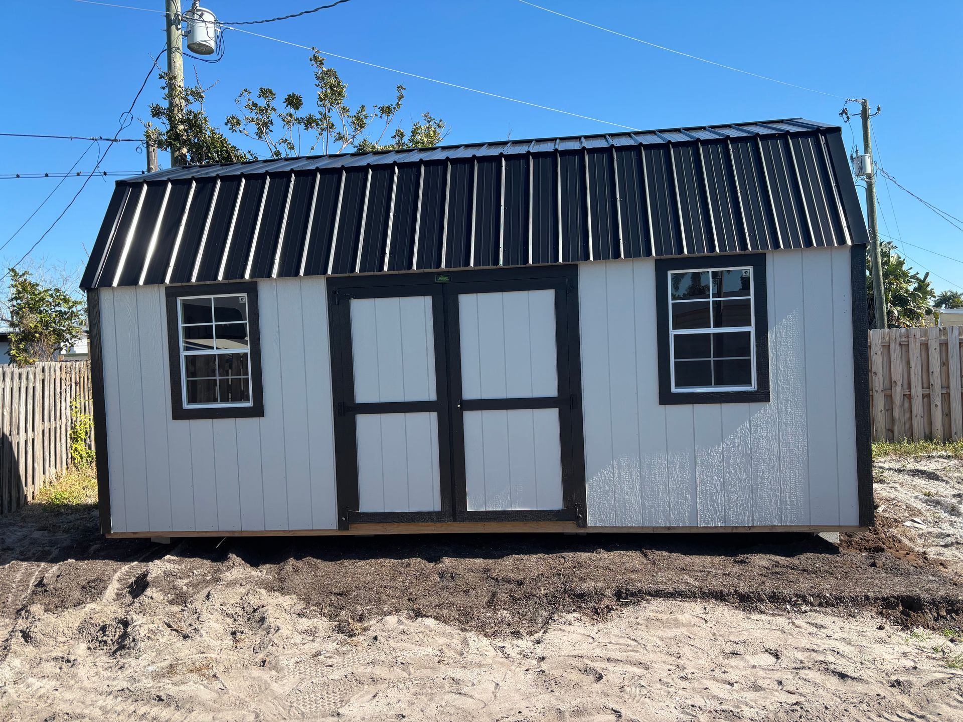 Small white shed with black roof and double doors, standing on bare ground outdoors.