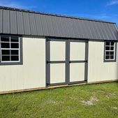 Small cream shed with dark roof, two windows, and double doors on a grassy lawn under a blue sky