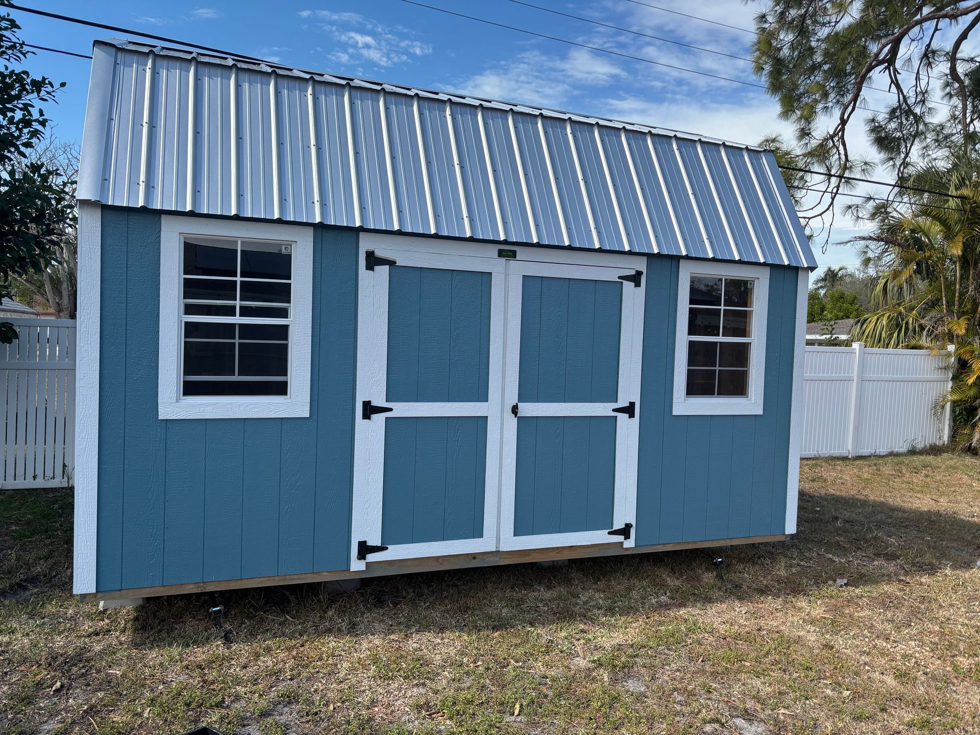Blue-and-white shed with double doors and two windows, set in a grassy yard under a partly cloudy sky