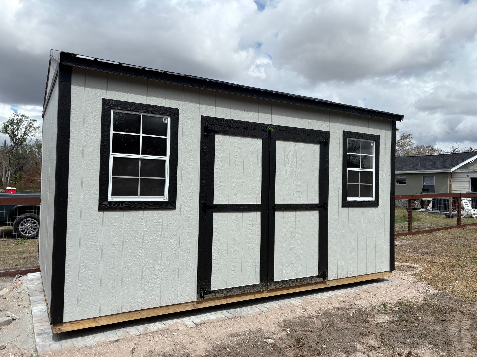 Small white shed with black trim and double doors on a dirt lot under a cloudy sky