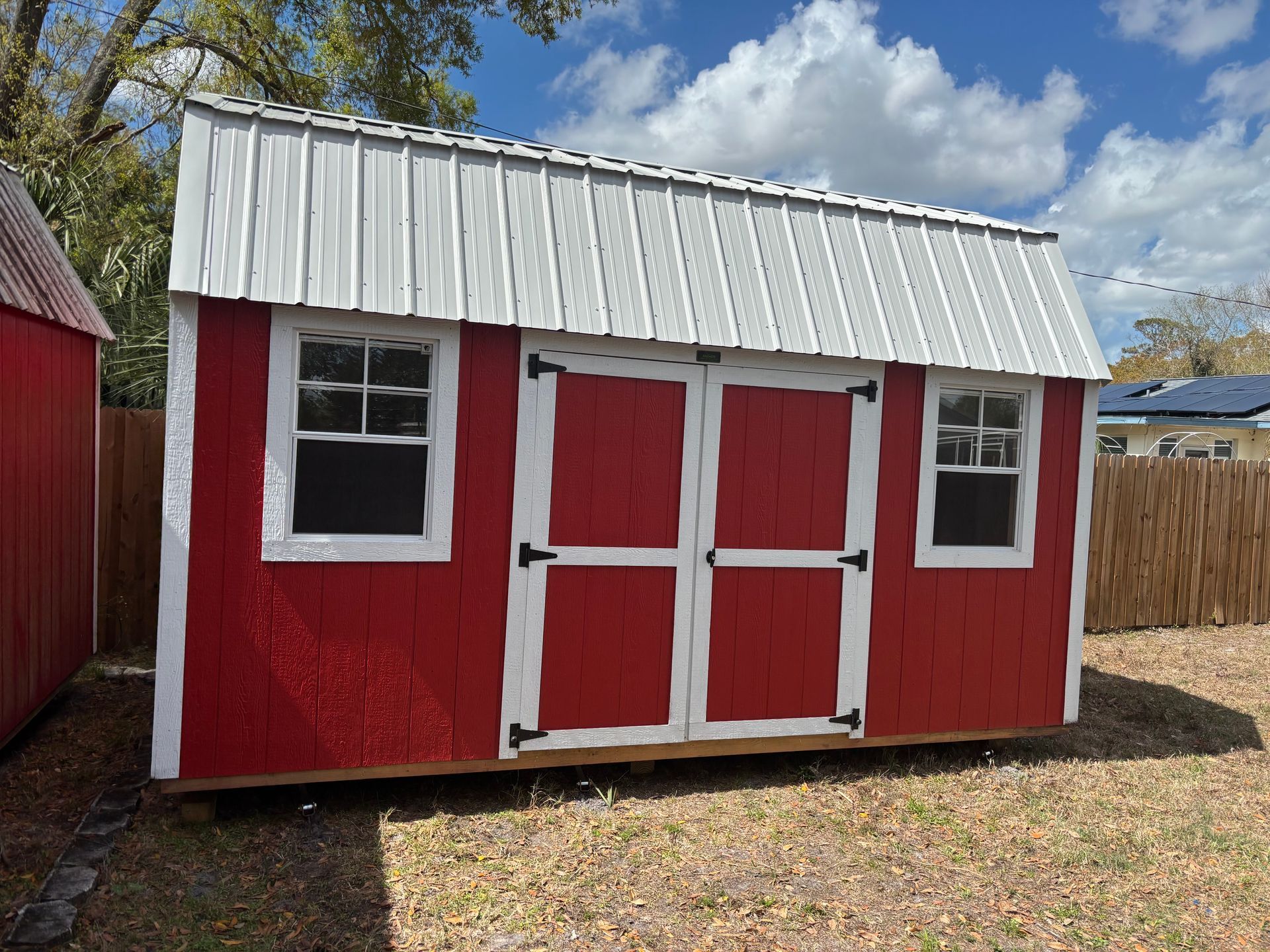 Red wooden shed with white trim and double doors in a backyard under a partly cloudy sky