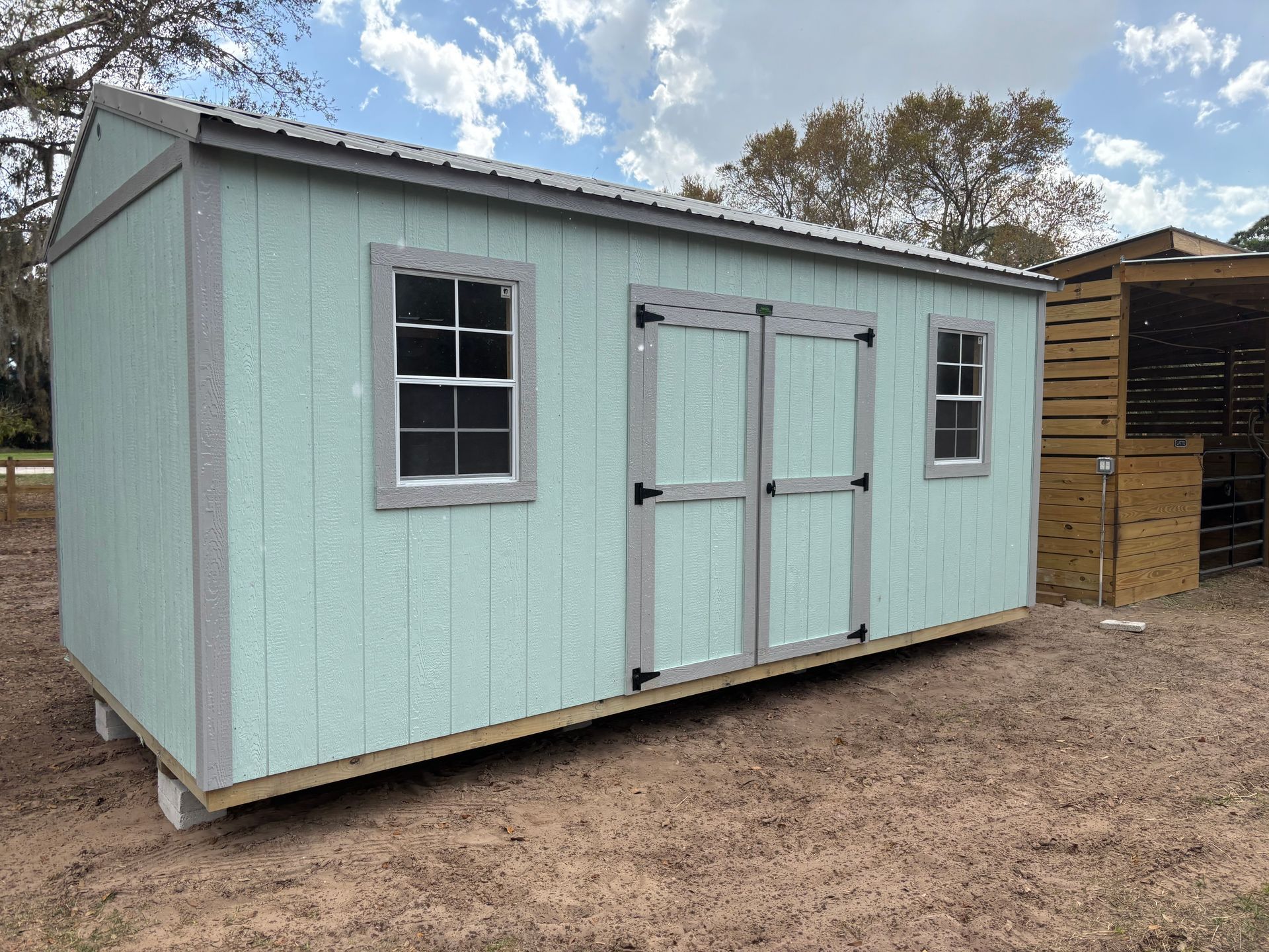 Mint-green storage shed with double doors and windows in a dirt yard