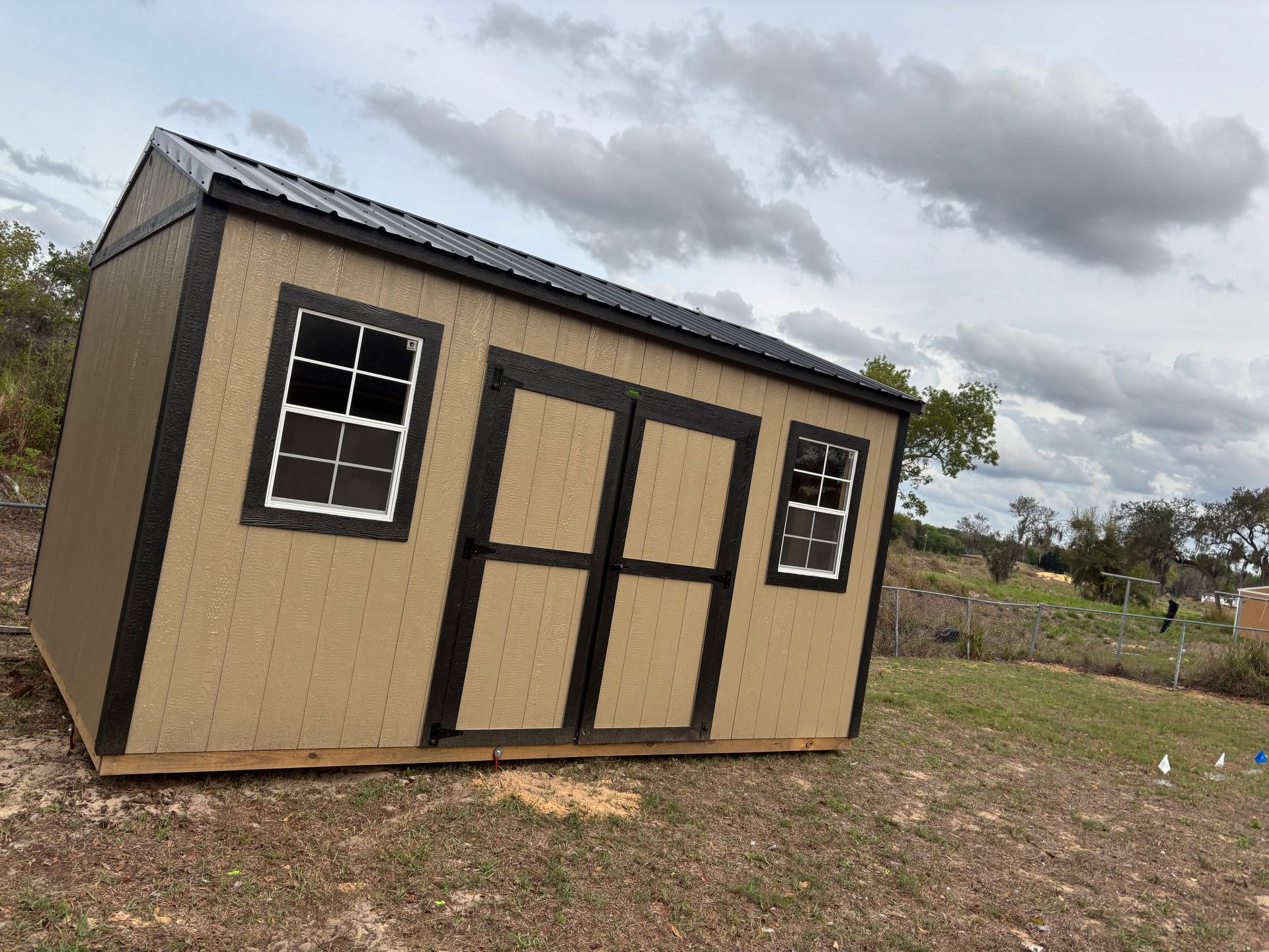 Small tan shed with black trim and windows, standing on a grassy lot under a cloudy sky.