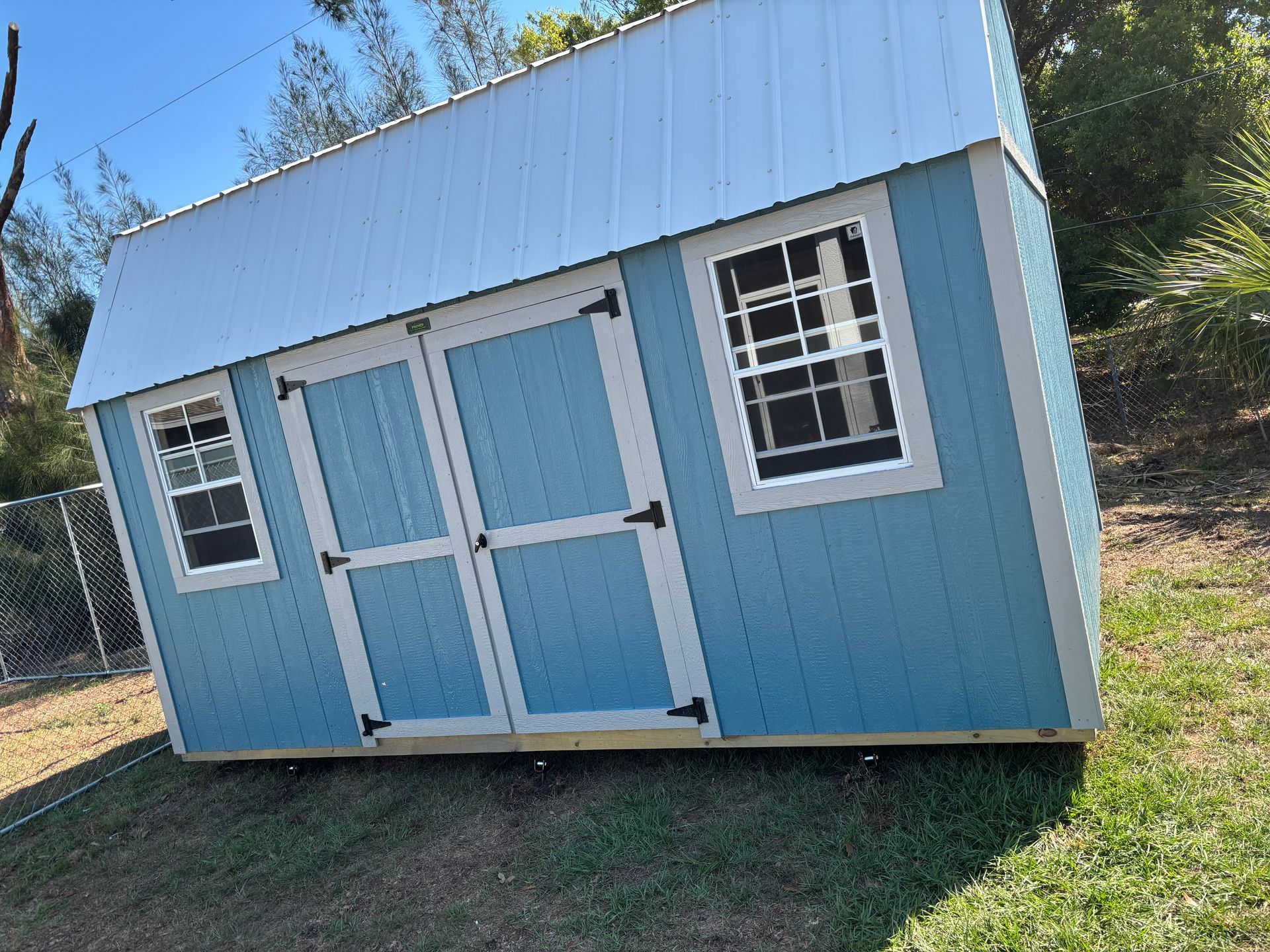 Light blue shed with white trim and double doors in a grassy yard, seen at an angle