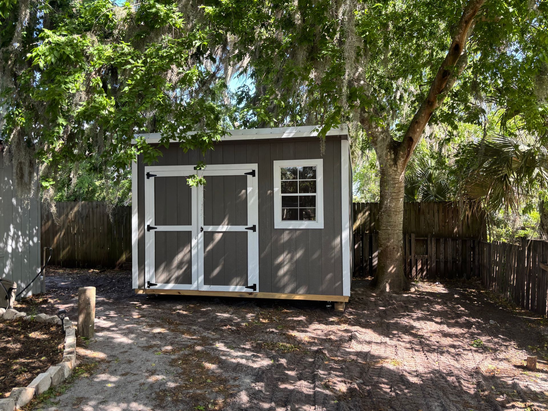 Small gray shed with double white doors and a window in a shaded backyard beneath trees