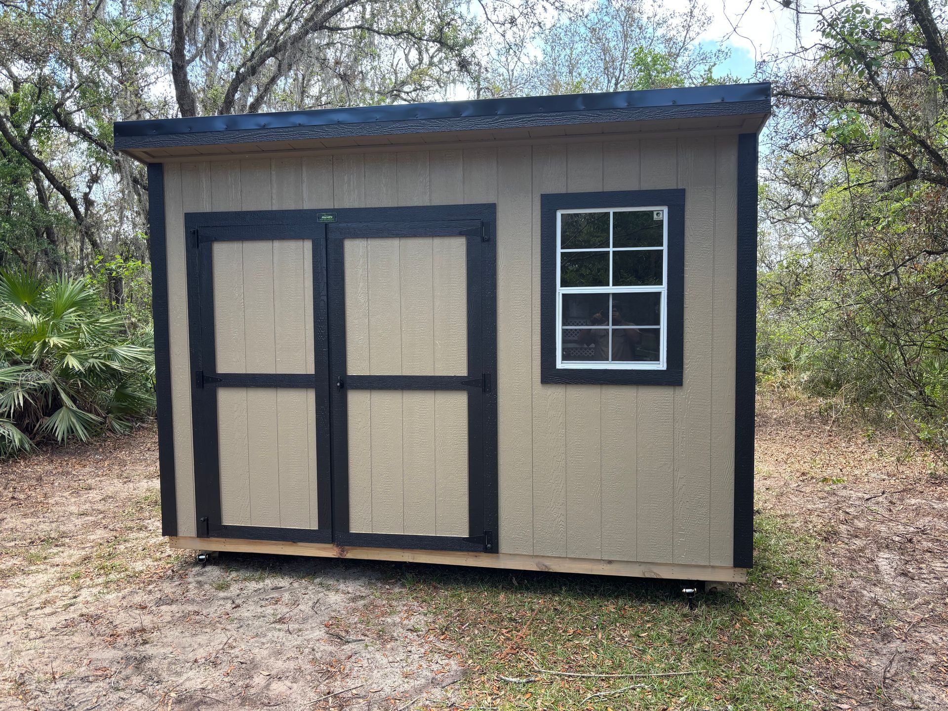 Small tan shed with dark trim, double doors, and a window in a wooded yard